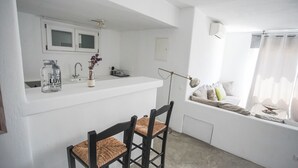 A small kitchenette with a white countertop, a sink, and a window with a view of the outside. There is a chair with a woven seat and a black frame. - Villa Carina by Paradise Estate Mykonos : Prime Location & Endless Sea Views (Platis Gialos, Mykonos)
