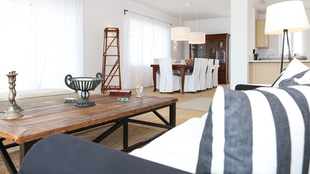 A living room with a wooden coffee table, striped pillows, a ladder, and a dining area in the background. - Villa Carina by Paradise Estate Mykonos : Prime Location & Endless Sea Views (Platis Gialos, Mykonos)