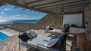 A sunlit outdoor seating area with stone walls, a pool, and a view of the sea. - Villa Carina by Paradise Estate Mykonos : Prime Location & Endless Sea Views (Platis Gialos, Mykonos)
