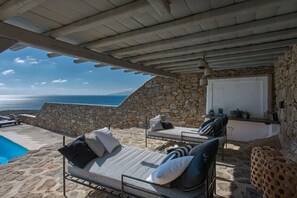 A sunlit outdoor seating area with stone walls, a pool, and a view of the sea. - Villa Carina by Paradise Estate Mykonos : Prime Location & Endless Sea Views (Platis Gialos, Mykonos)