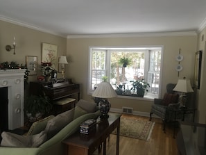 A living room with a piano, sofa, and a large window with plants.