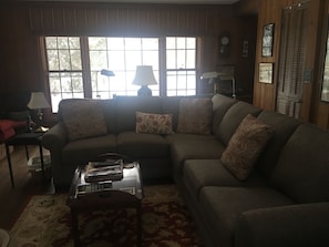 A living room with a sectional sofa, a coffee table, and a patterned rug. There is a window with blinds, a lamp, and a clock on the wall.