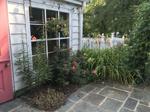 A garden with a variety of plants and flowers, a white picket fence, and a stone pathway leading to a house with a pink door.