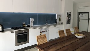 A modern kitchen with white cabinets, a blue backsplash, a gas stove, a dishwasher, and a wooden dining table with wicker chairs. - One of best locations at Coast. Surrounded by nature and the sound of surf. (Adelaide)