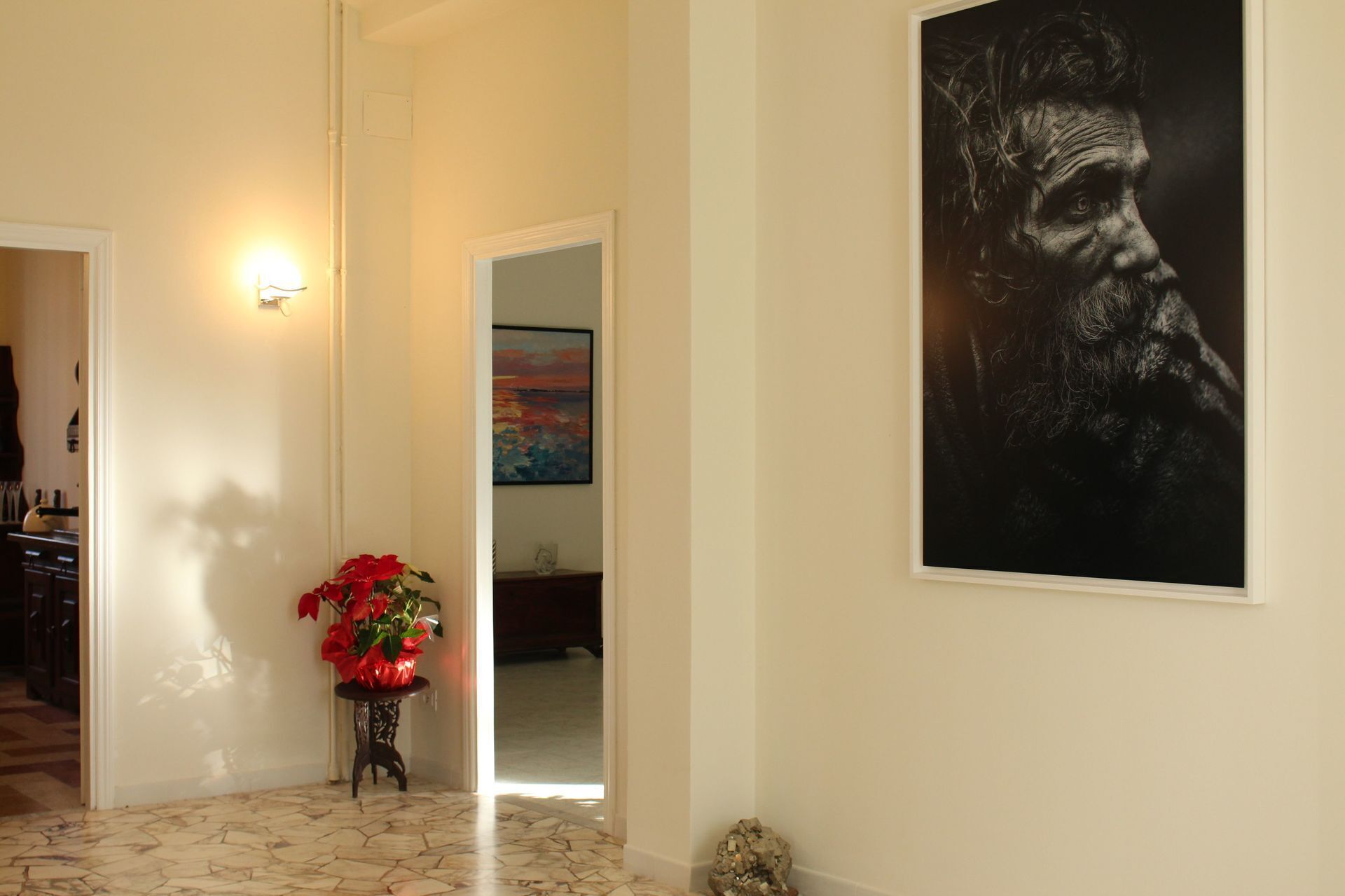 A hallway with a tiled floor, a wall-mounted light, a framed painting, and a potted plant.