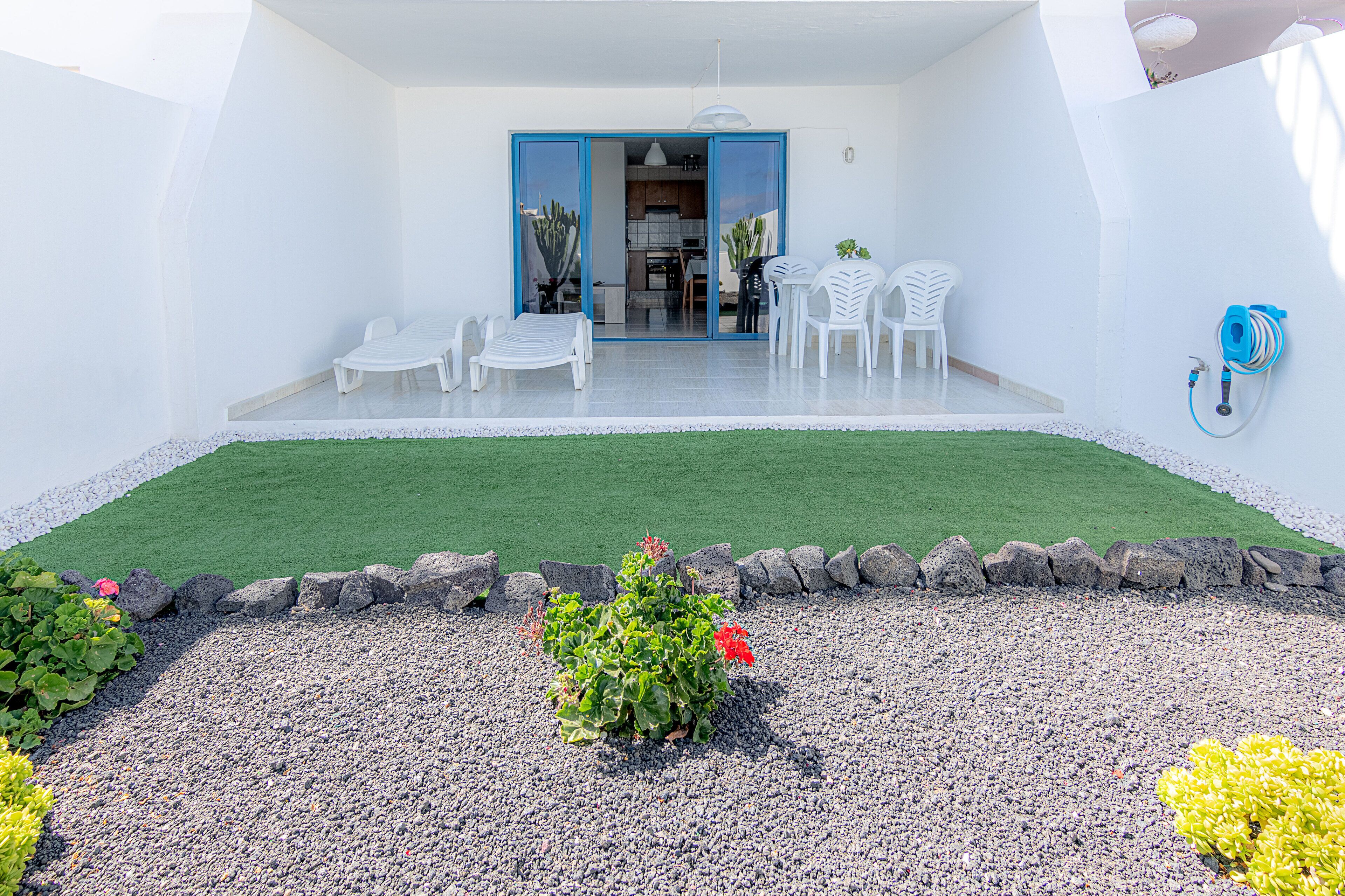 Une terrasse avec des chaises blanches et une table, un petit jardin agrémenté de rochers et de plantes, et une porte bleue donnant accès à un espace intérieur.
