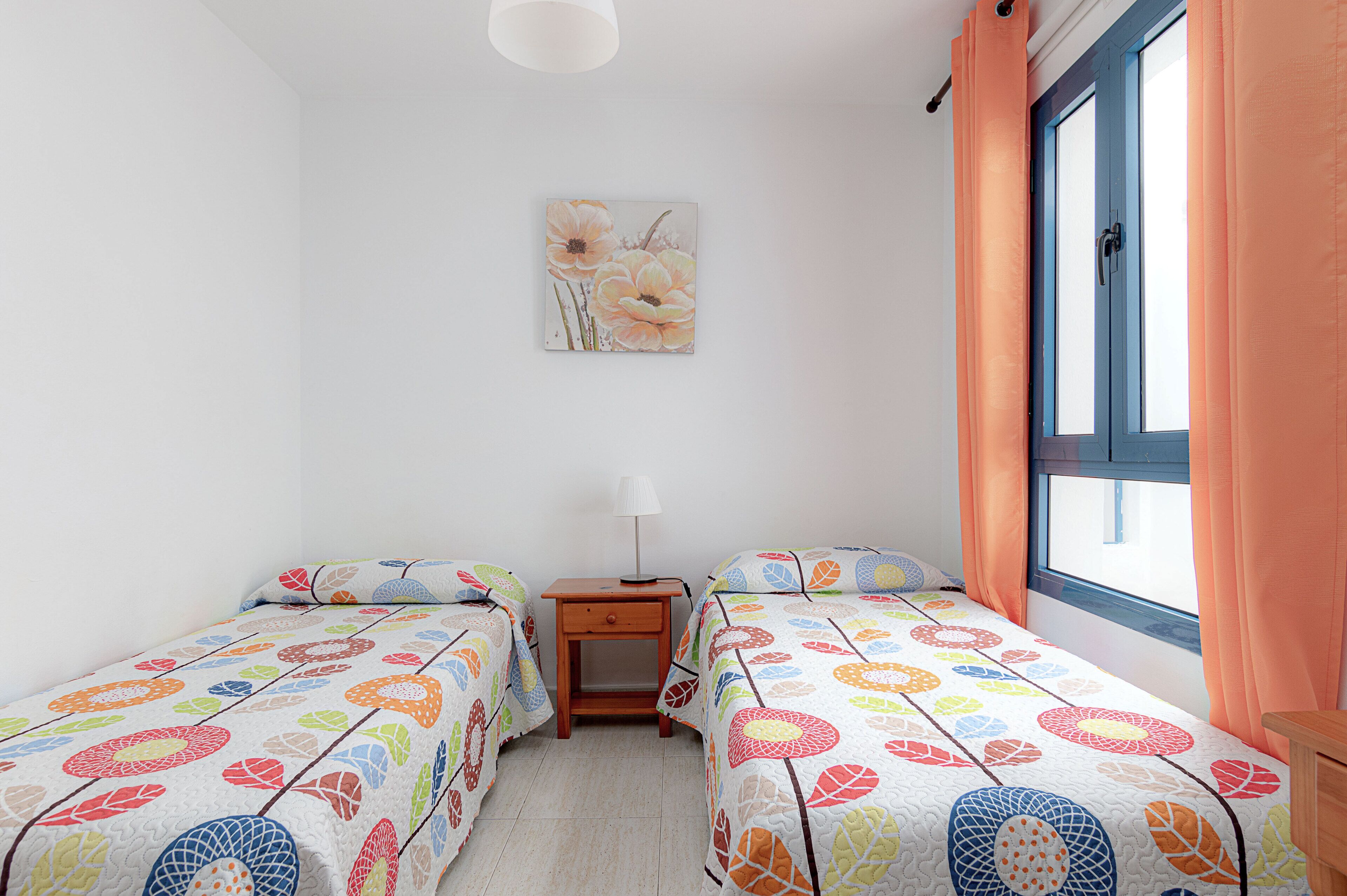 A bedroom with two single beds, a nightstand, a lamp, and a window with curtains.
