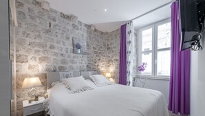 Une chambre avec un mur en pierre, un lit avec une literie blanche, deux tables de chevet avec des lampes, des rideaux violets et une fenêtre donnant sur un beau paysage.