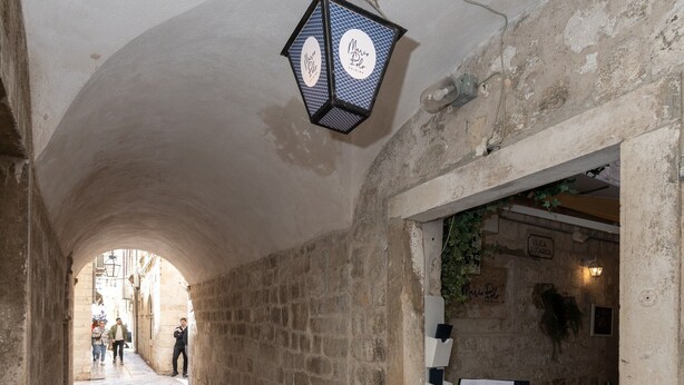 Un étroit couloir en pierre, éclairé par une lanterne suspendue, avec des murs de pierre et une table entourée de chaises, le tout situé sur le côté droit.