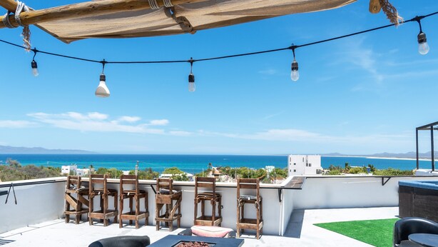 Terraza en la azotea con sillas de madera, una mesa y luces colgantes, con cielo azul despejado y una costa lejana al fondo.