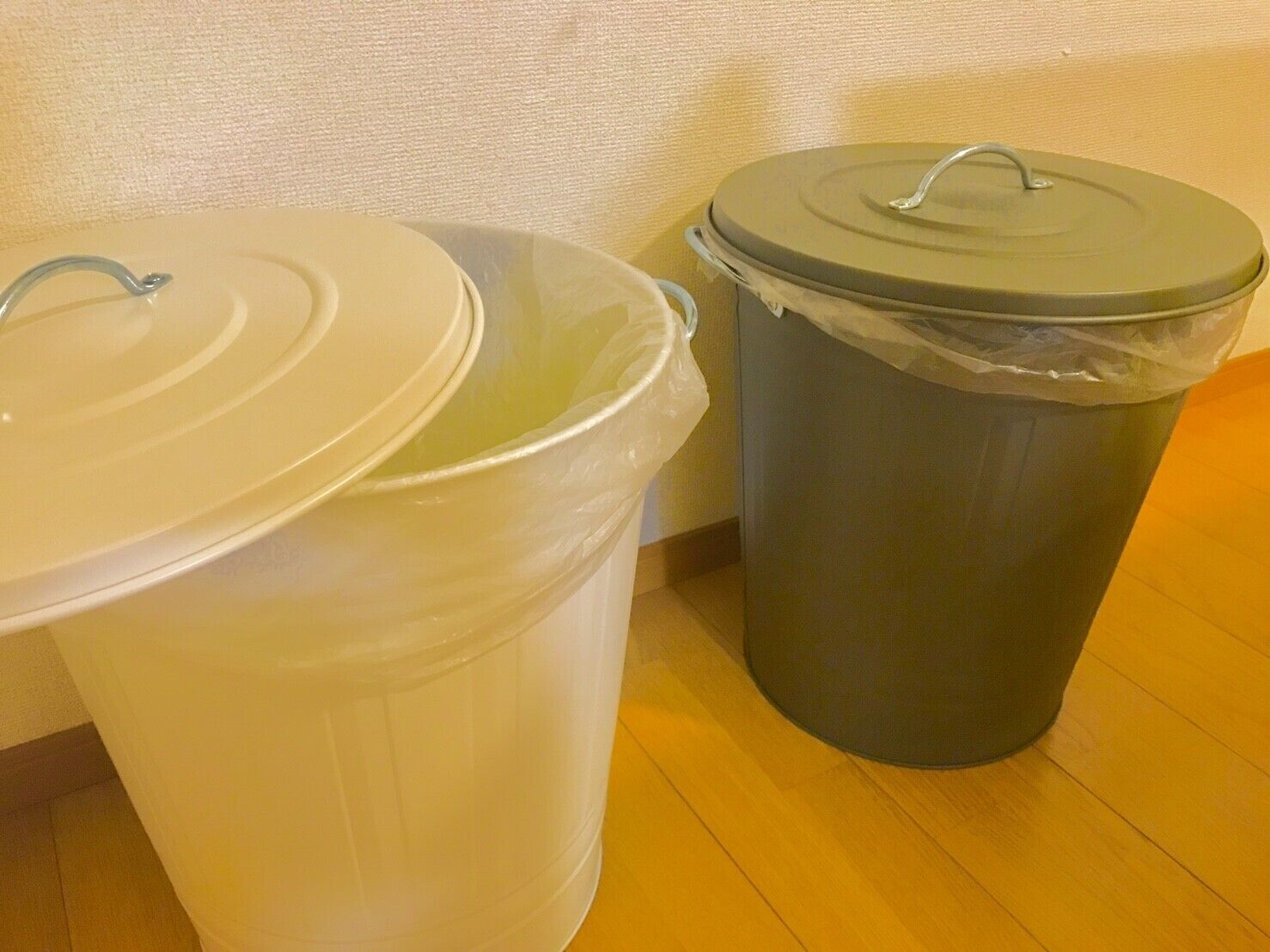 Two trash bins, one white and one gray, with lids, placed on a wooden floor against a textured wall.