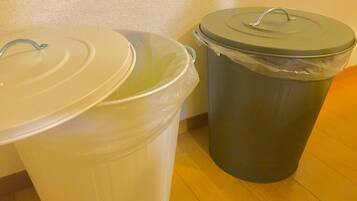 Two trash bins, one white and one gray, with lids, placed on a wooden floor against a textured wall.