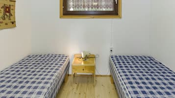 Pieni huone, jossa on kaksi erillistä sänkyä, puinen yöpöytä lampun kanssa ja ikkuna koristeellisella kehyksellä.