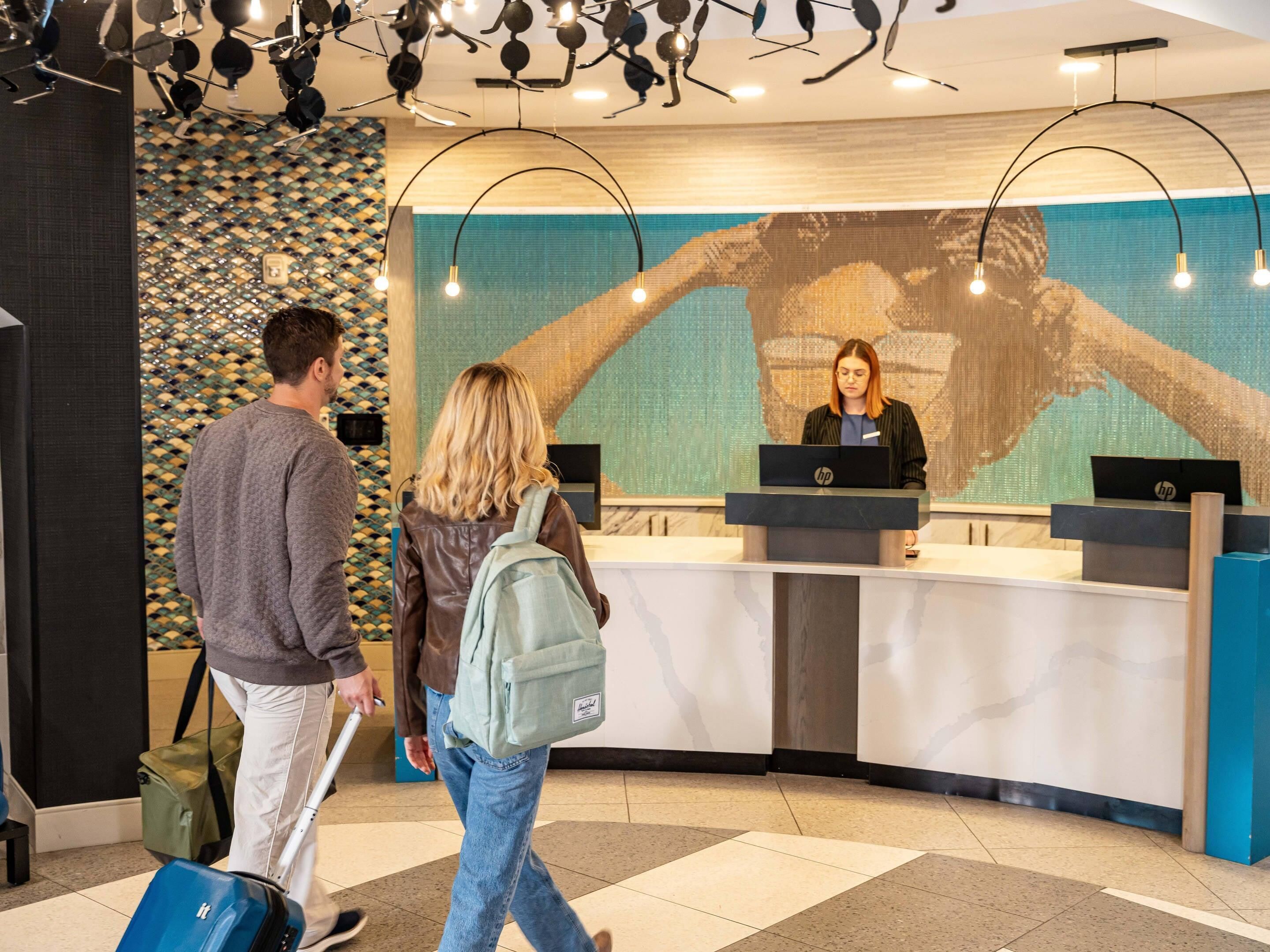 A reception desk with a large digital screen displaying a handprint, a man with a suitcase, and a woman with a backpack.