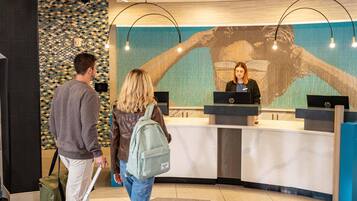A reception desk with a large digital screen displaying a handprint, a man with a suitcase, and a woman with a backpack.