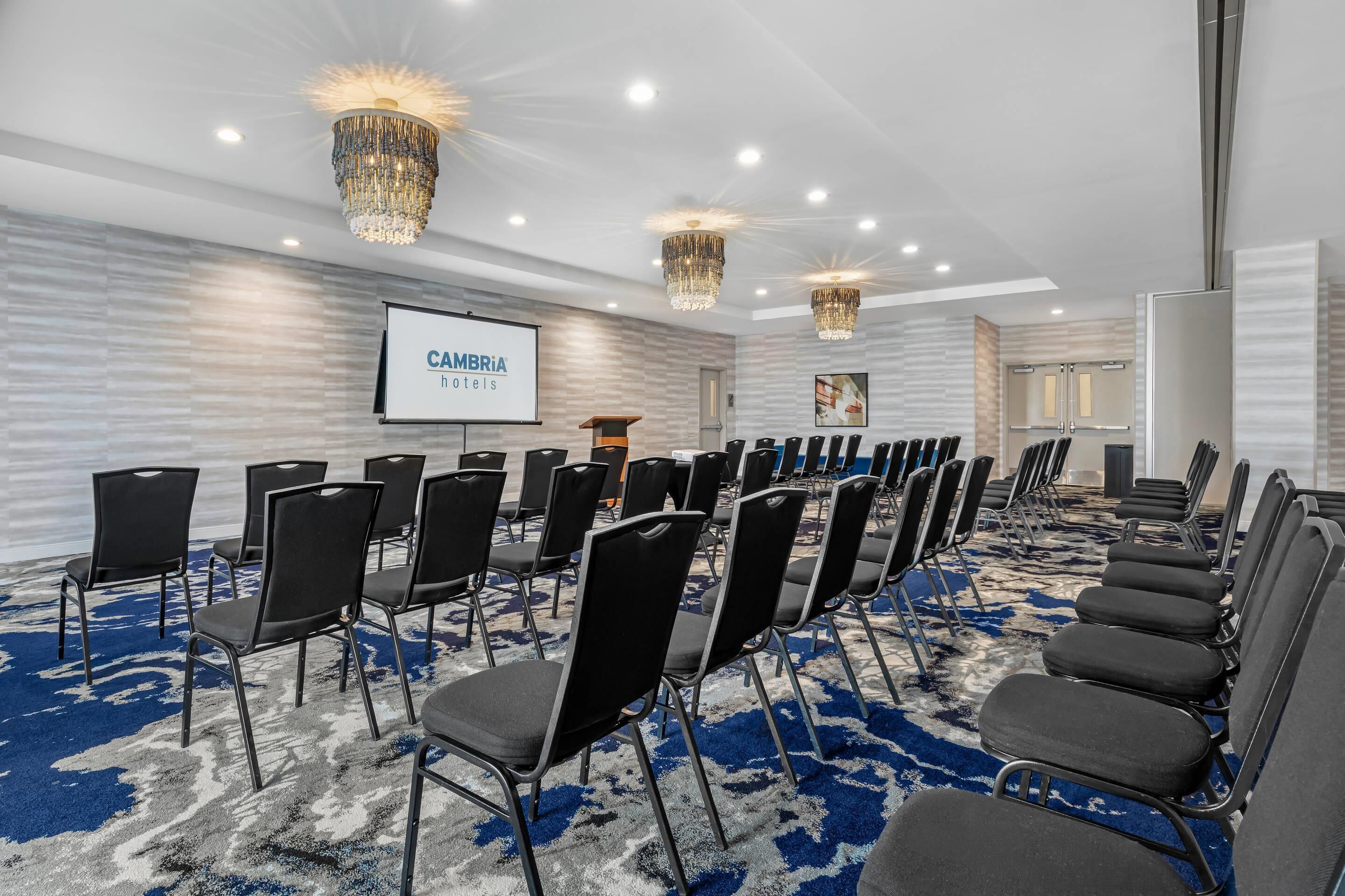 Un salón de conferencias con filas de sillas negras, una pantalla de proyector y candelabros.