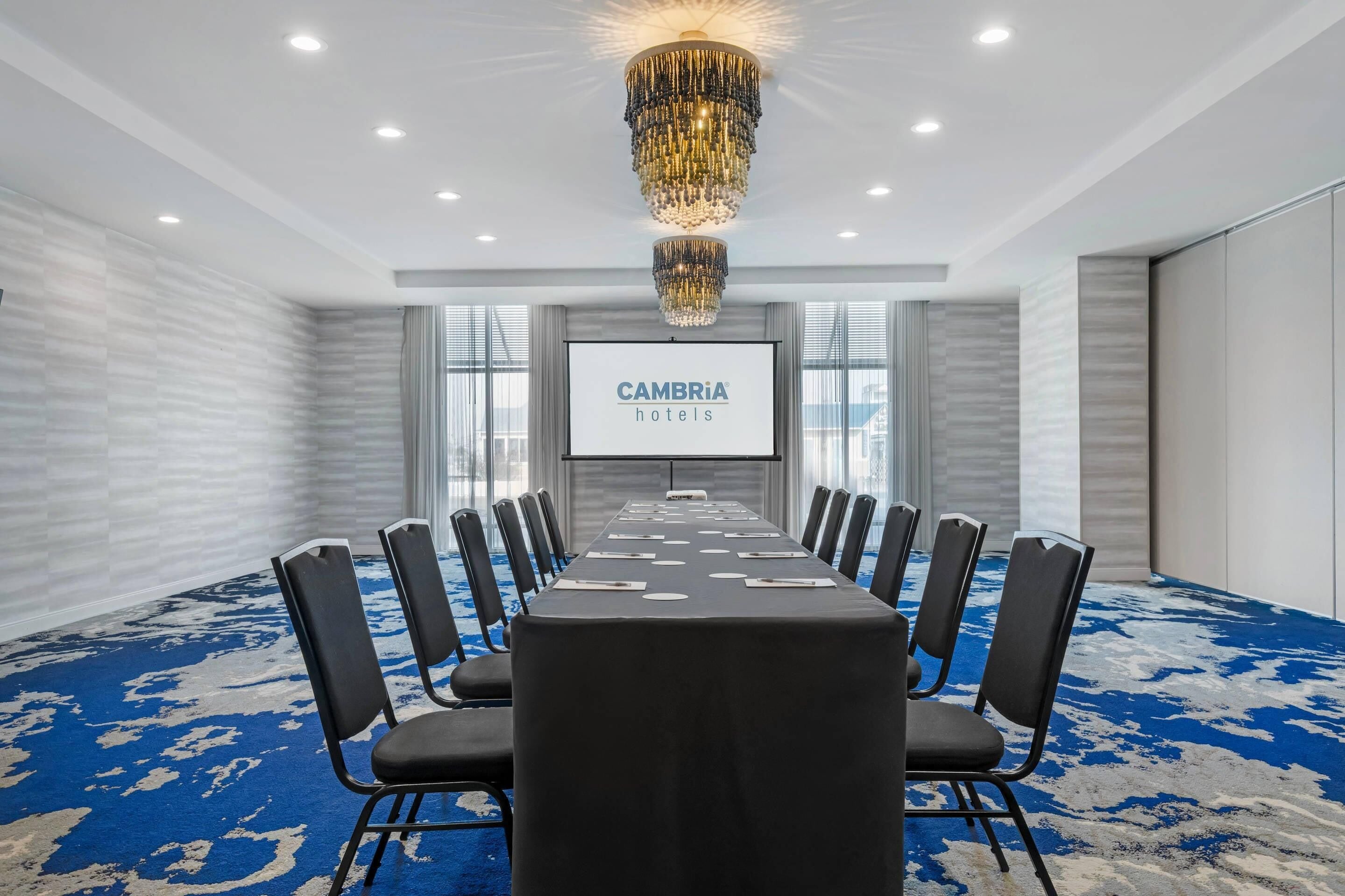 A conference room with a long table, black chairs, a chandelier, and a screen displaying 'CAMBRIA hotels'.