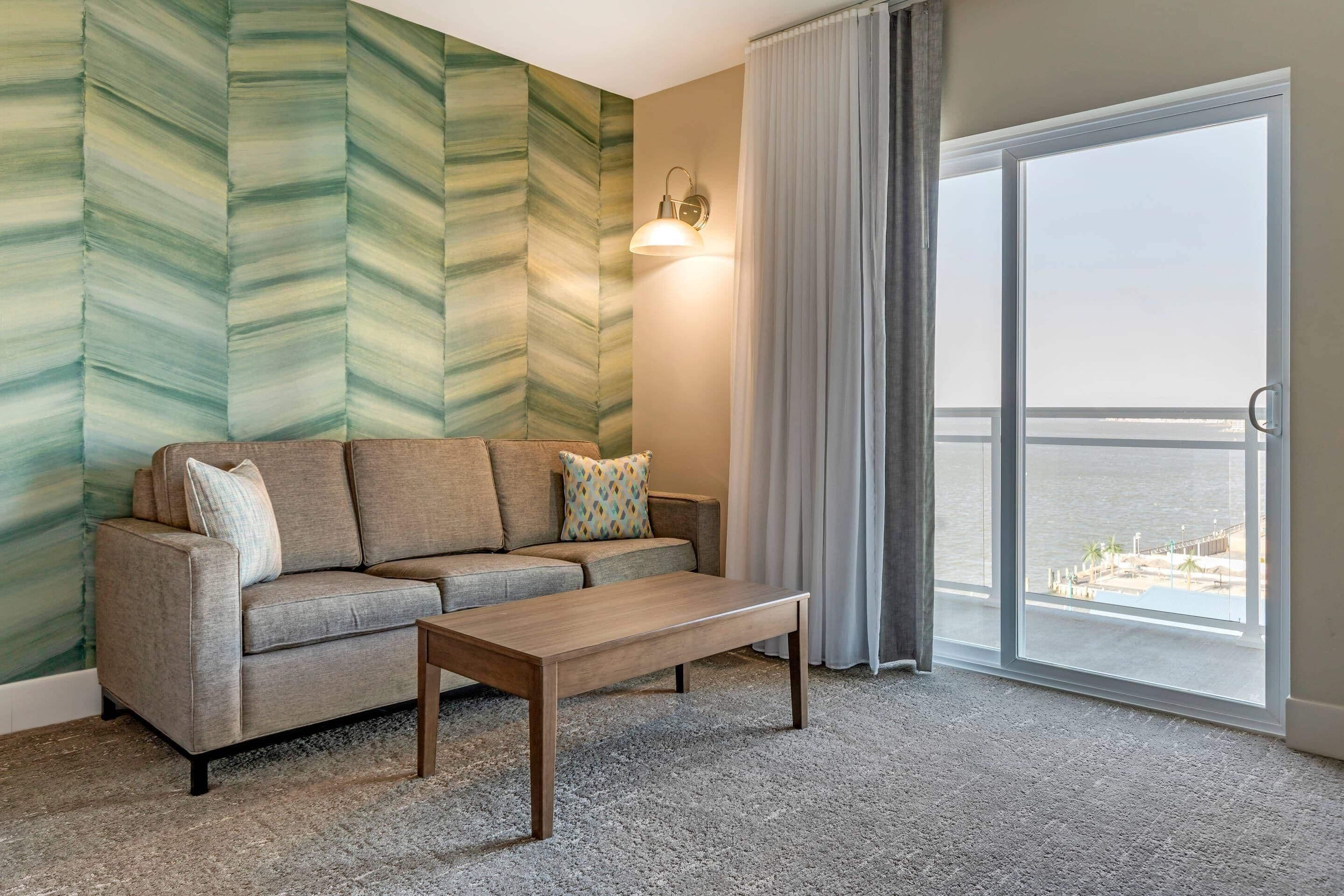 A room with a sofa, a wooden coffee table, a wall with a green pattern, a window with a view of the sea, and a light fixture.