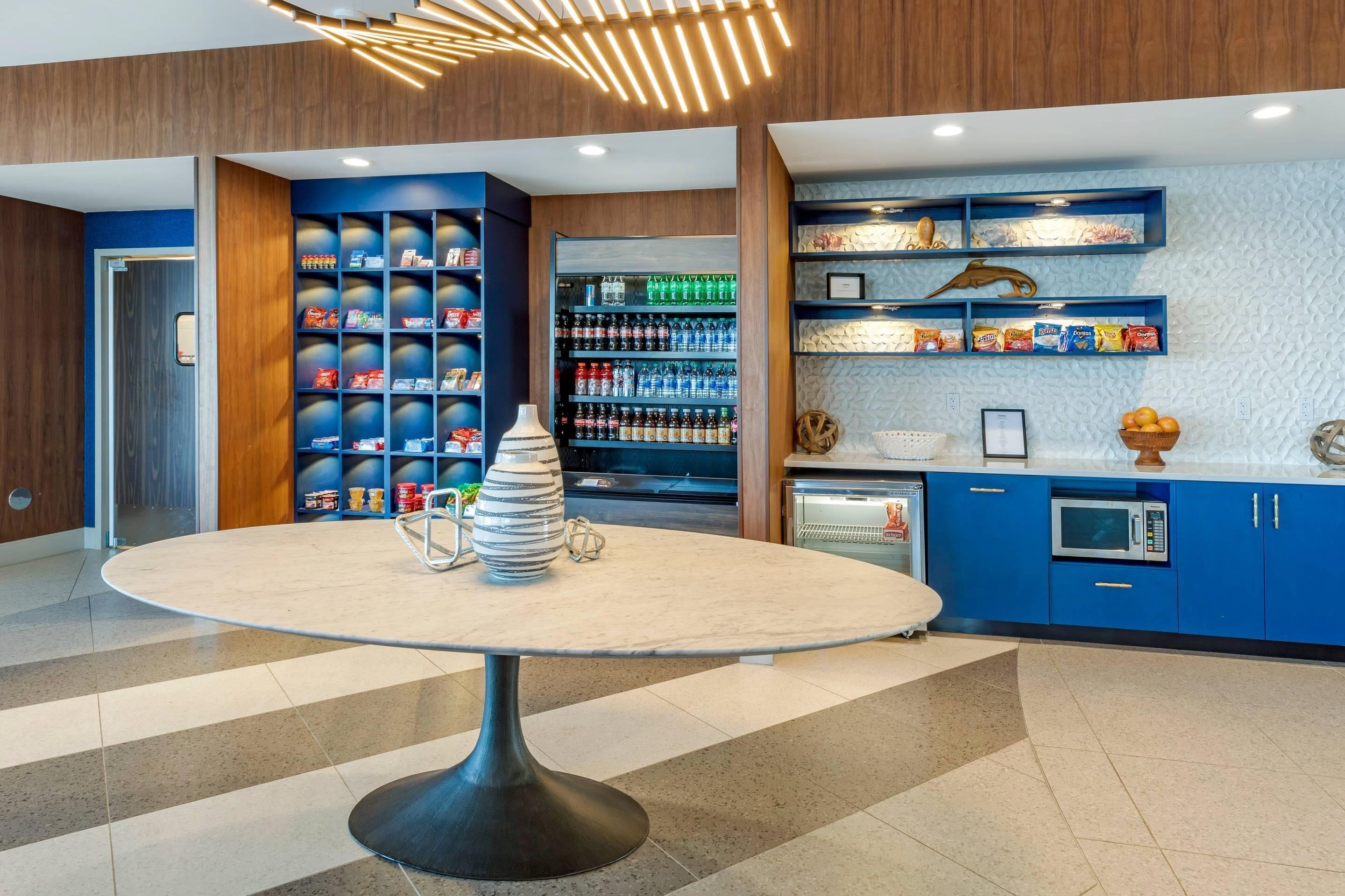 A modern interior with a round table, shelves with snacks, a microwave, and a refrigerator.