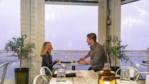 A dining area with a table set for two, potted plants, and a view through large windows. - Cambria Hotel Ocean City - Bayfront (Ocean City)