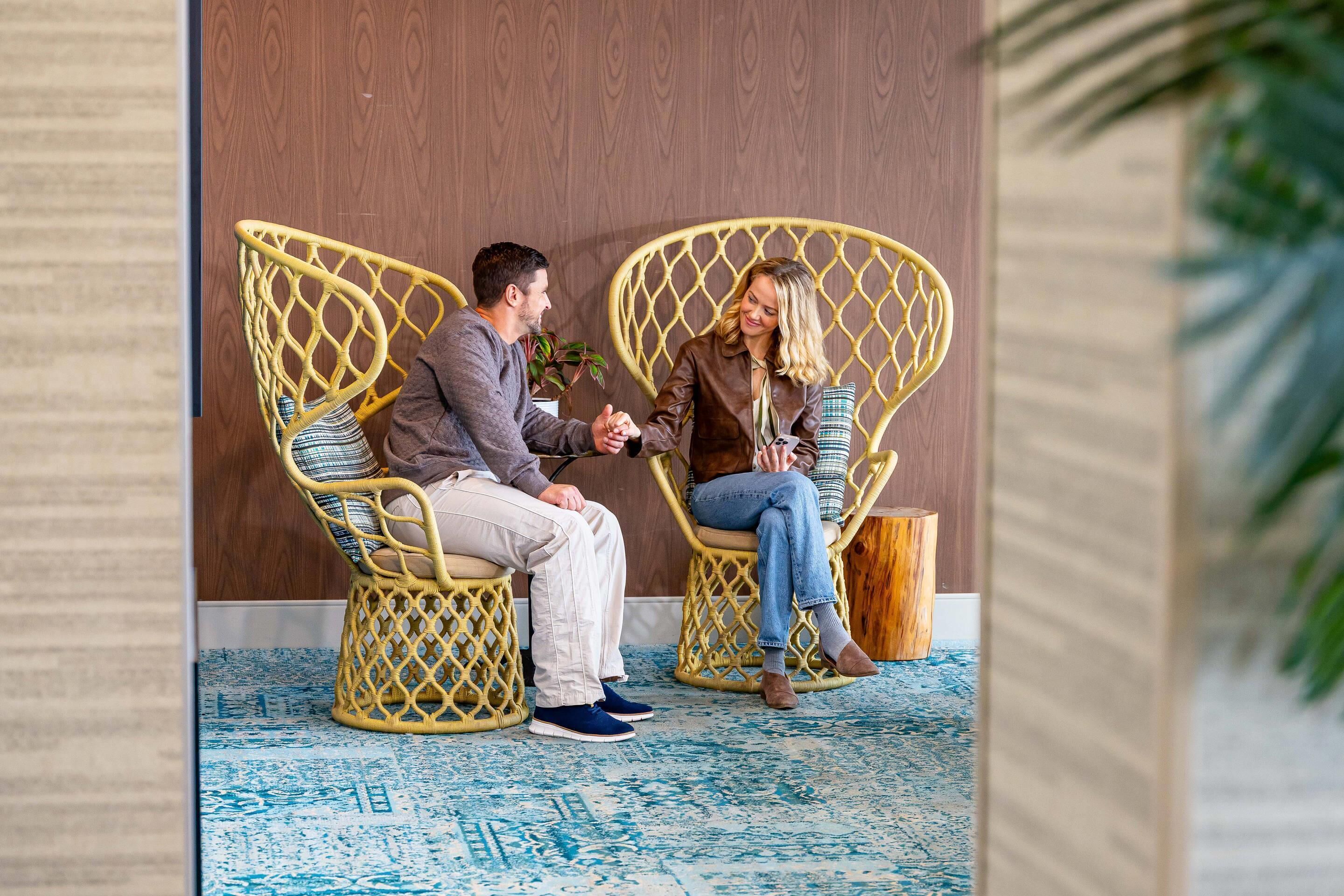 Two people sitting on wicker chairs in a room with a wooden wall and a blue patterned floor.