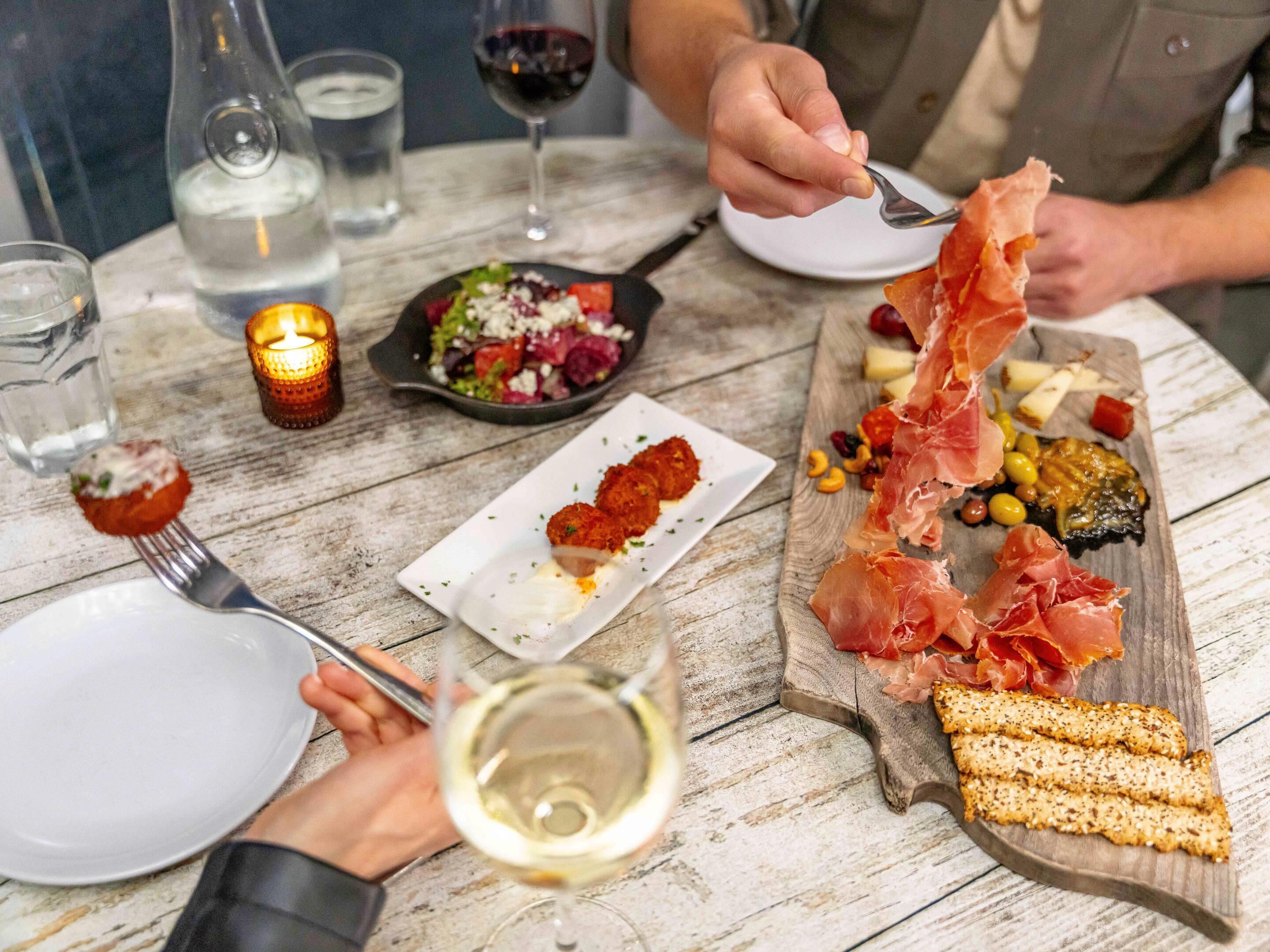 A dining table with various dishes, including a plate of prosciutto, a bowl of salad, and a glass of white wine.