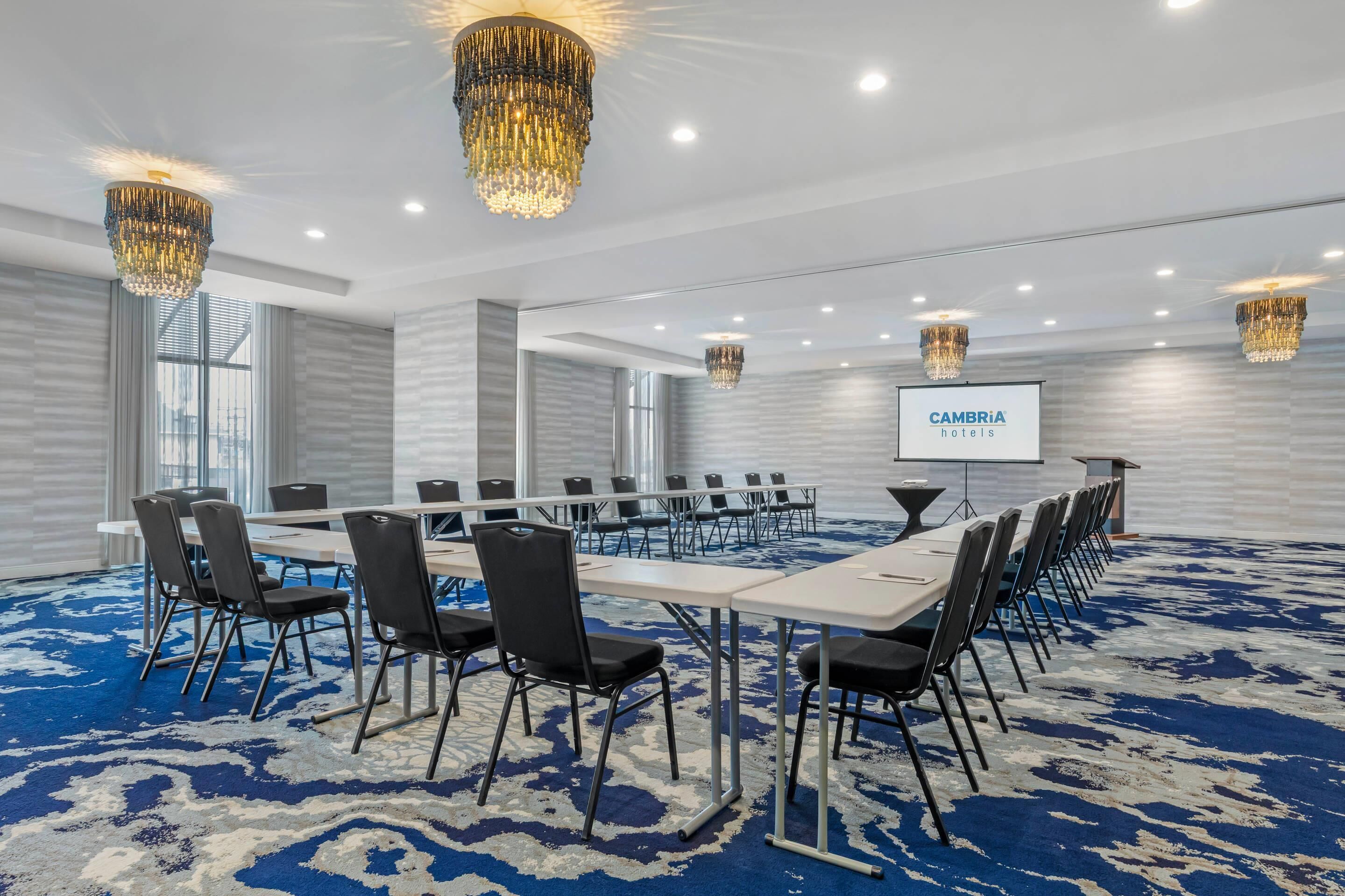 A conference room with a long table, chairs, chandeliers, and a screen displaying 'CAMBRIA hotels'.