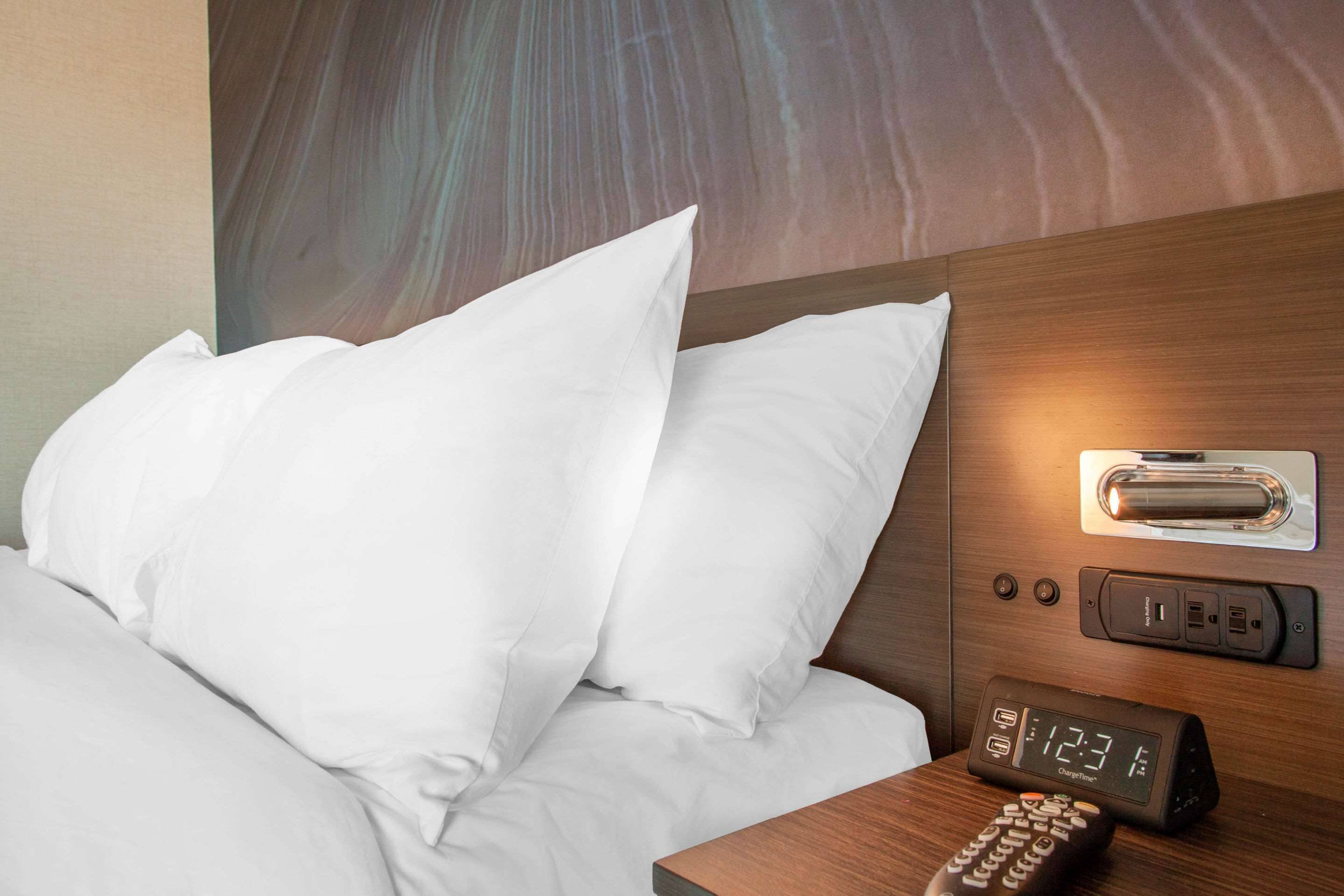 A hotel room with a bed, white pillows, a wooden headboard, a bedside table with a remote control, and a digital clock displaying the time.