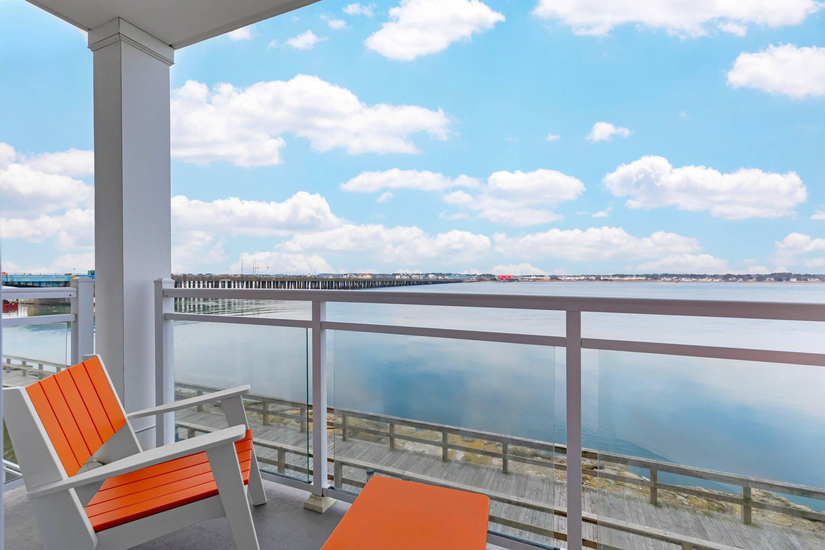 A balcony with a view of the water, a wooden pier, and two orange chairs.