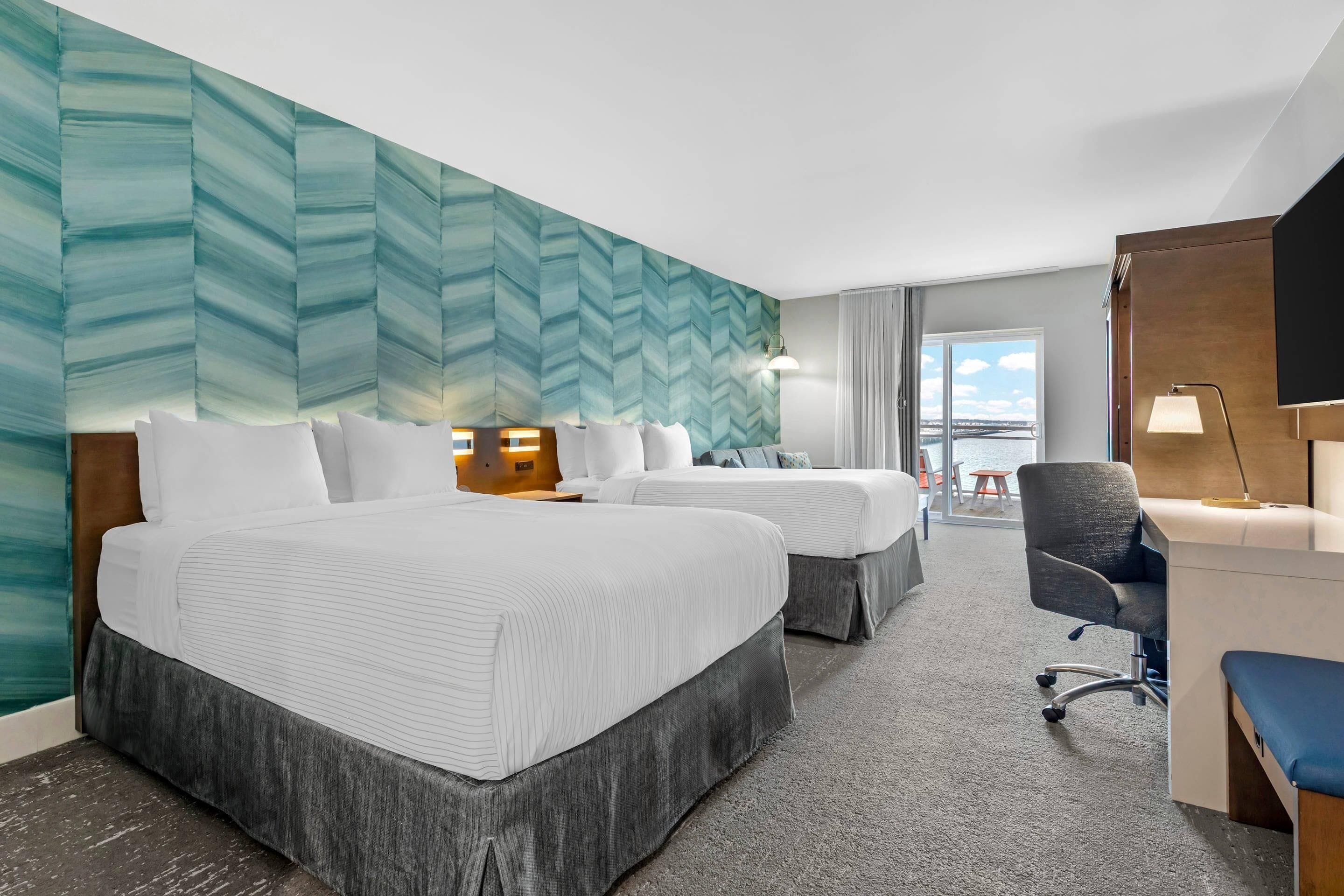 A hotel room with two beds, a desk, a chair, and a view of the sky.