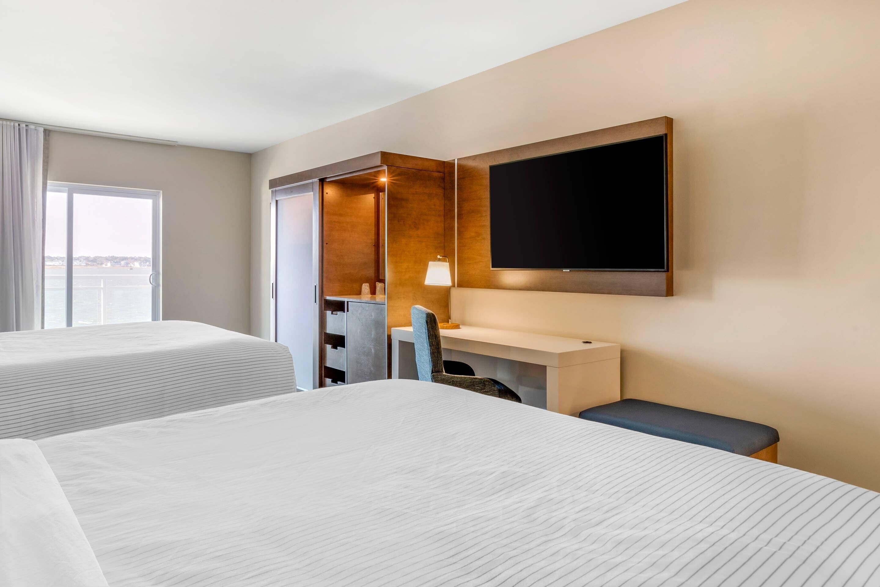 A hotel room with two beds, a desk, a chair, a TV, and a view of the outside.