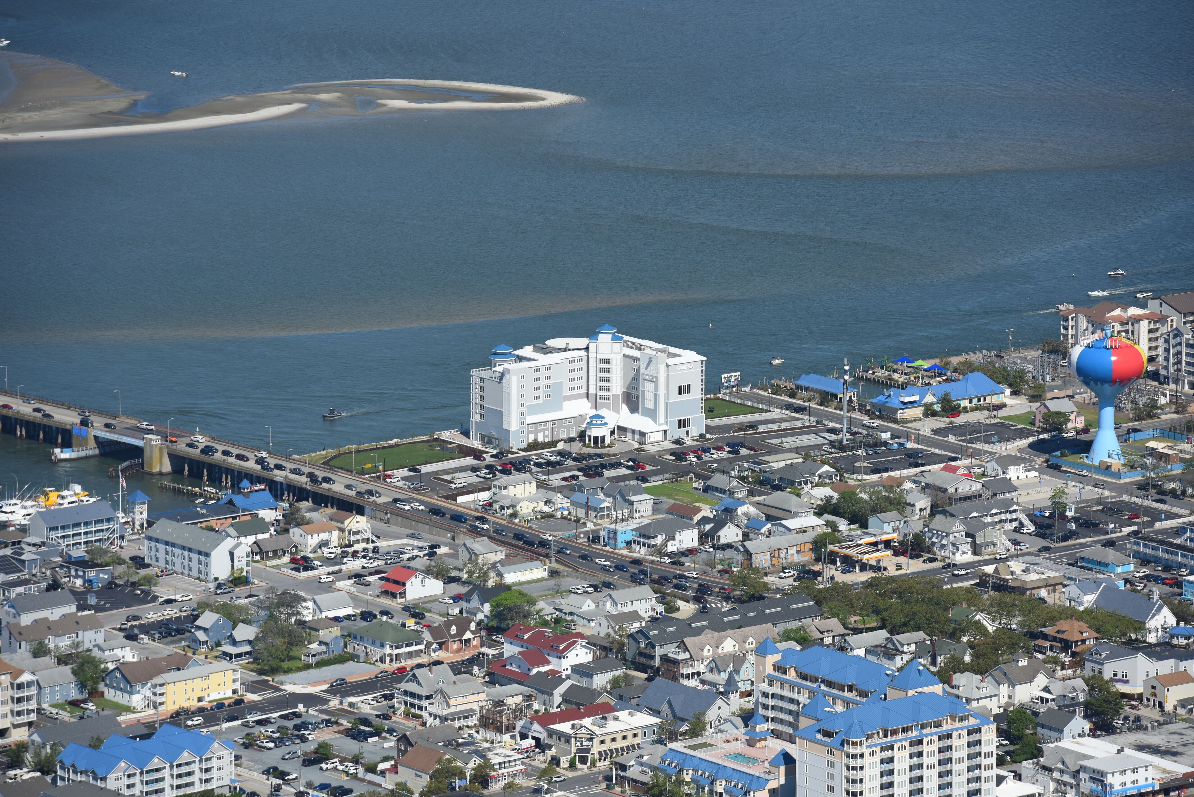 Vista aérea de un pueblo costero con un edificio blanco destacado de techo azul, una torre de agua azul y roja, y un puente que lo conecta a tierra firme.