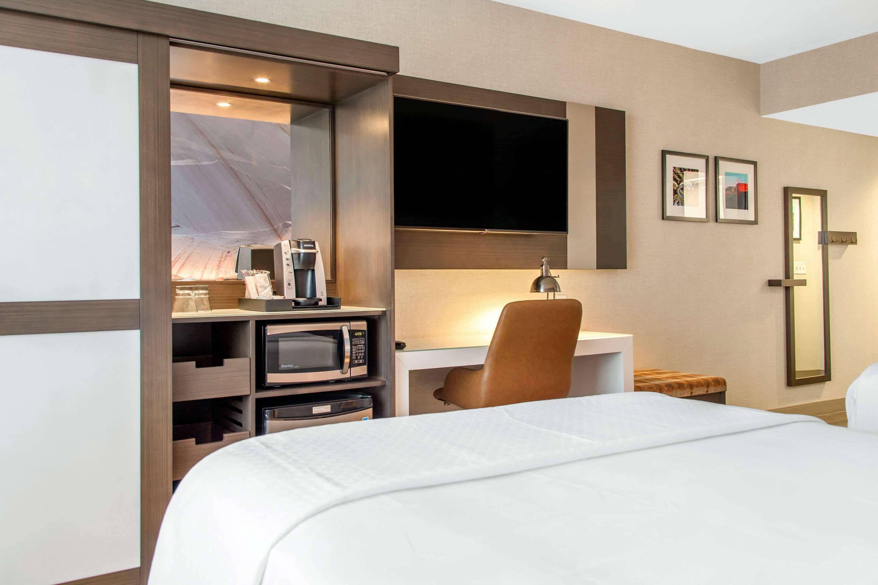 A hotel room with a bed, a desk with a computer, a microwave, a coffee maker, and a TV mounted on the wall.