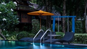 Un espace piscine avec des chaises longues et un parasol, entouré d’une végétation luxuriante et d’un bâtiment en bois.