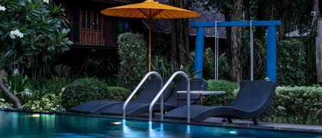 A pool area with lounge chairs and an umbrella, surrounded by lush greenery and a wooden building.