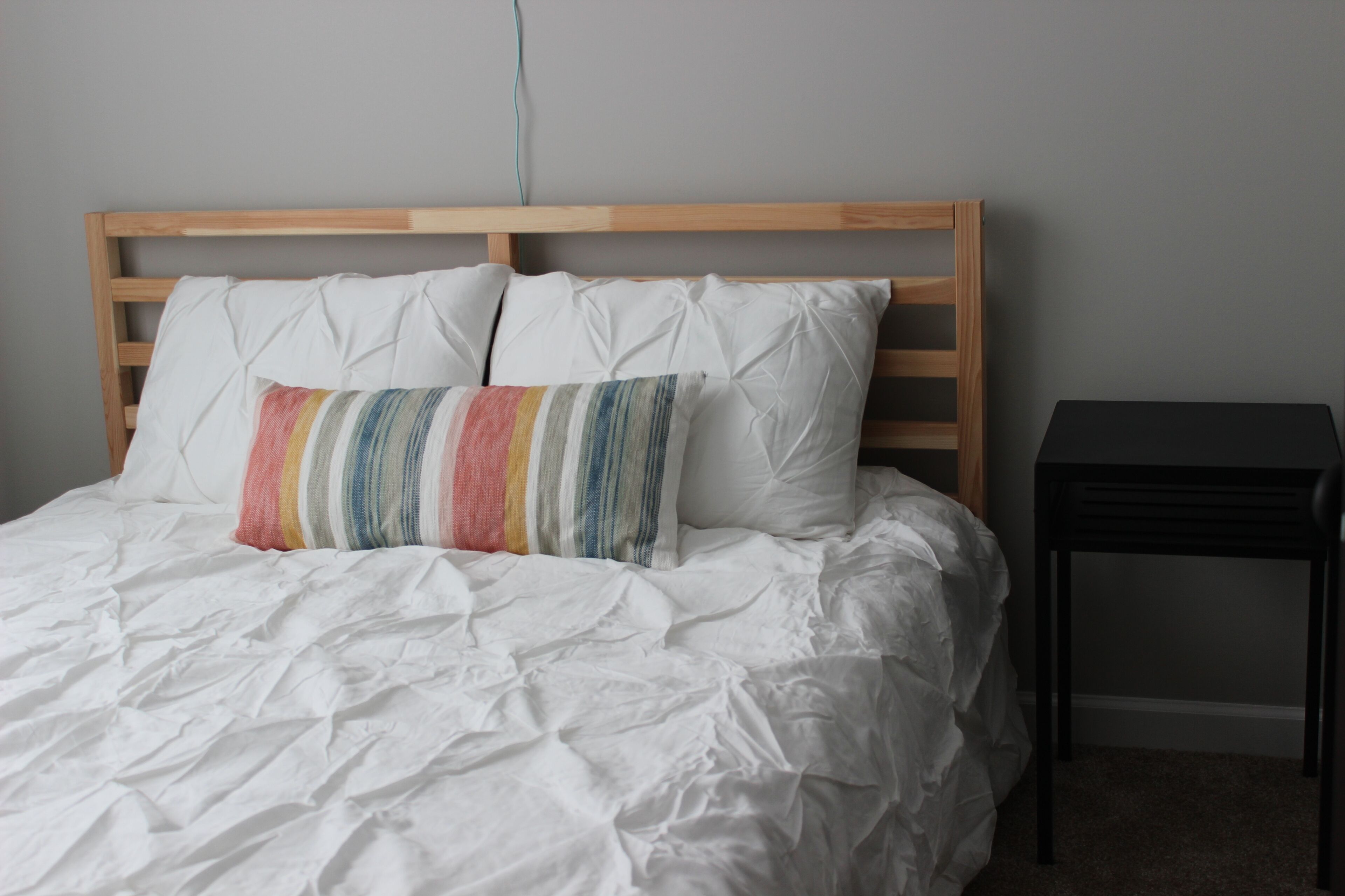 Una cama con sábanas blancas, un cojín a rayas de colores, un cabecero de madera y una mesita de noche negra.