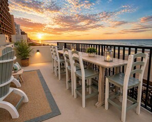 A terrace with a wooden table and chairs, a potted plant, and a view of the sunset over the ocean.