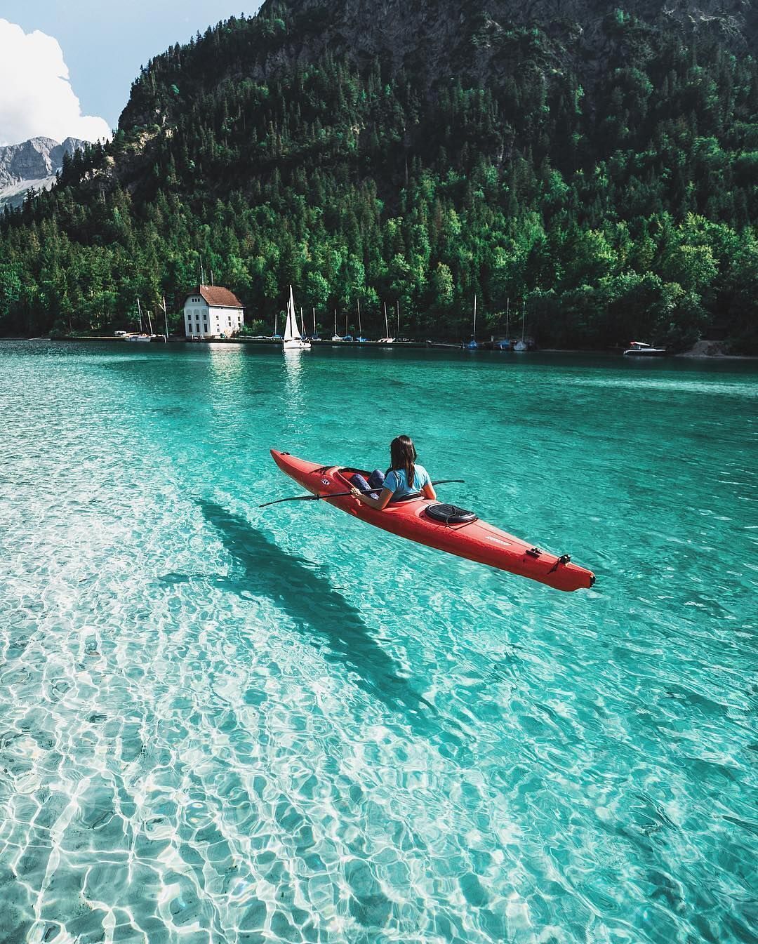 A kayaker in a red kayak on clear turquoise water with a house and sailboats in the background.