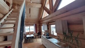 Interior de una cabaña de madera con escalera, cocina, comedor y ventanas amplias.