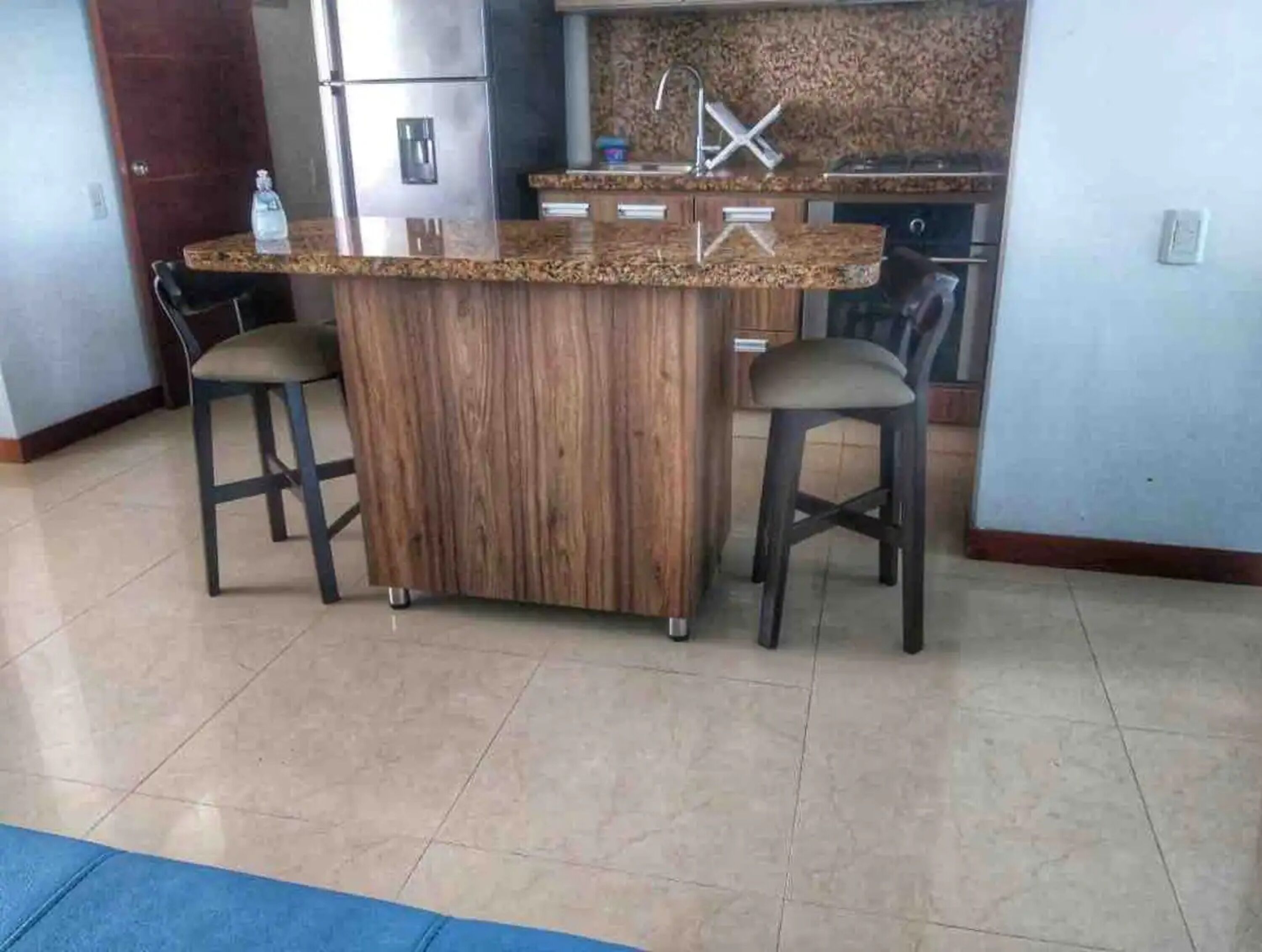 Una cocina con una isla central, dos taburetes de bar, un refrigerador y un lavamanos.