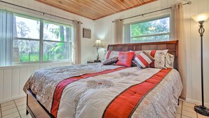 Un dormitorio con techo de madera, ventanas amplias, cama con ropa de cama estampada y una lámpara de noche.