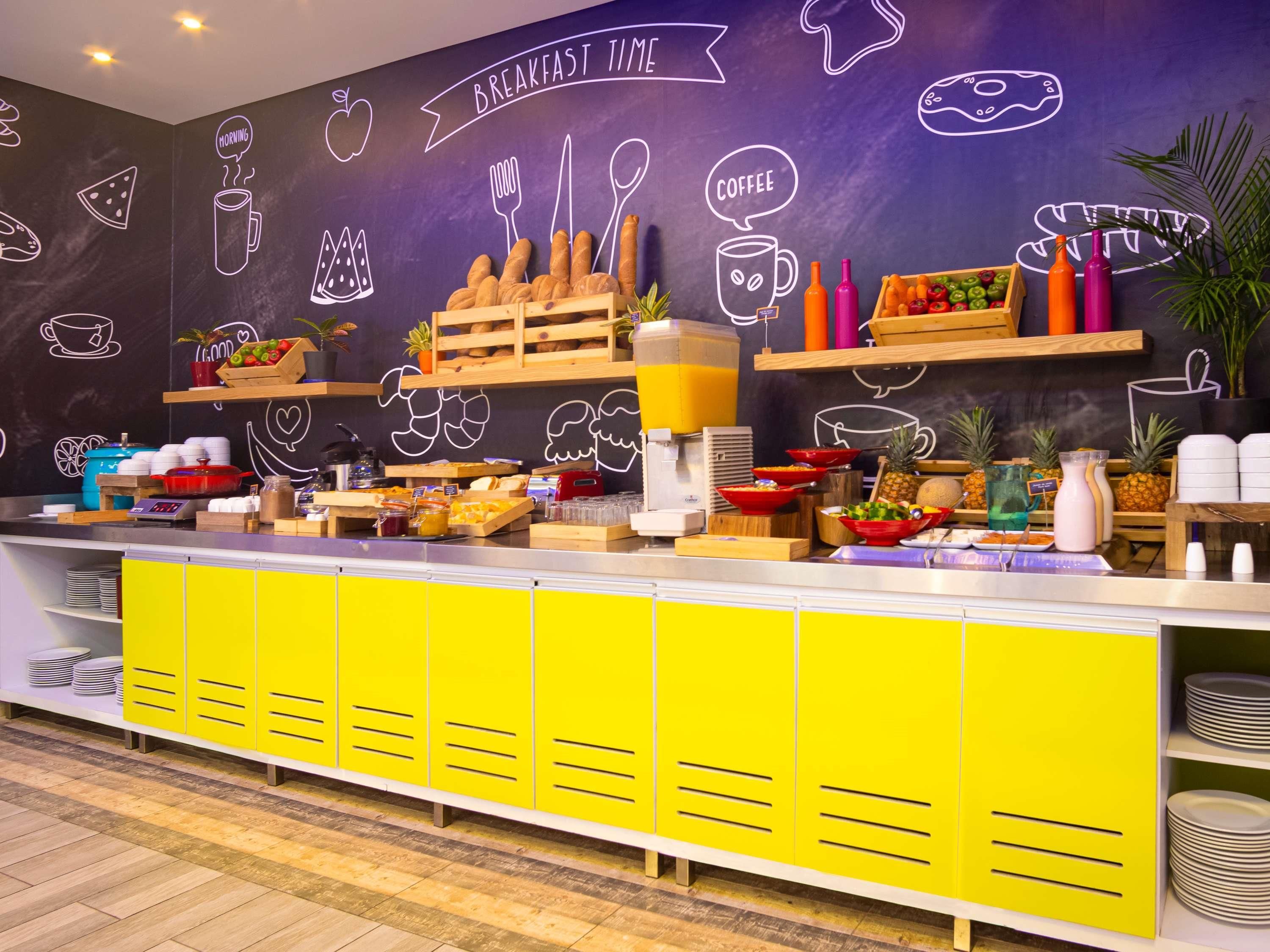 Un bufé de desayuno con diversas opciones de comida y bebidas, una pared de pizarra con dibujos relacionados con la comida, y gabinetes de color amarillo brillante.