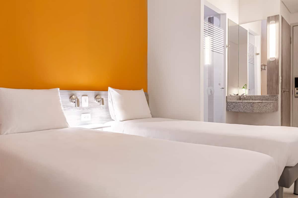 A hotel room with two beds, a bright orange accent wall, a large mirror, and a granite countertop.
