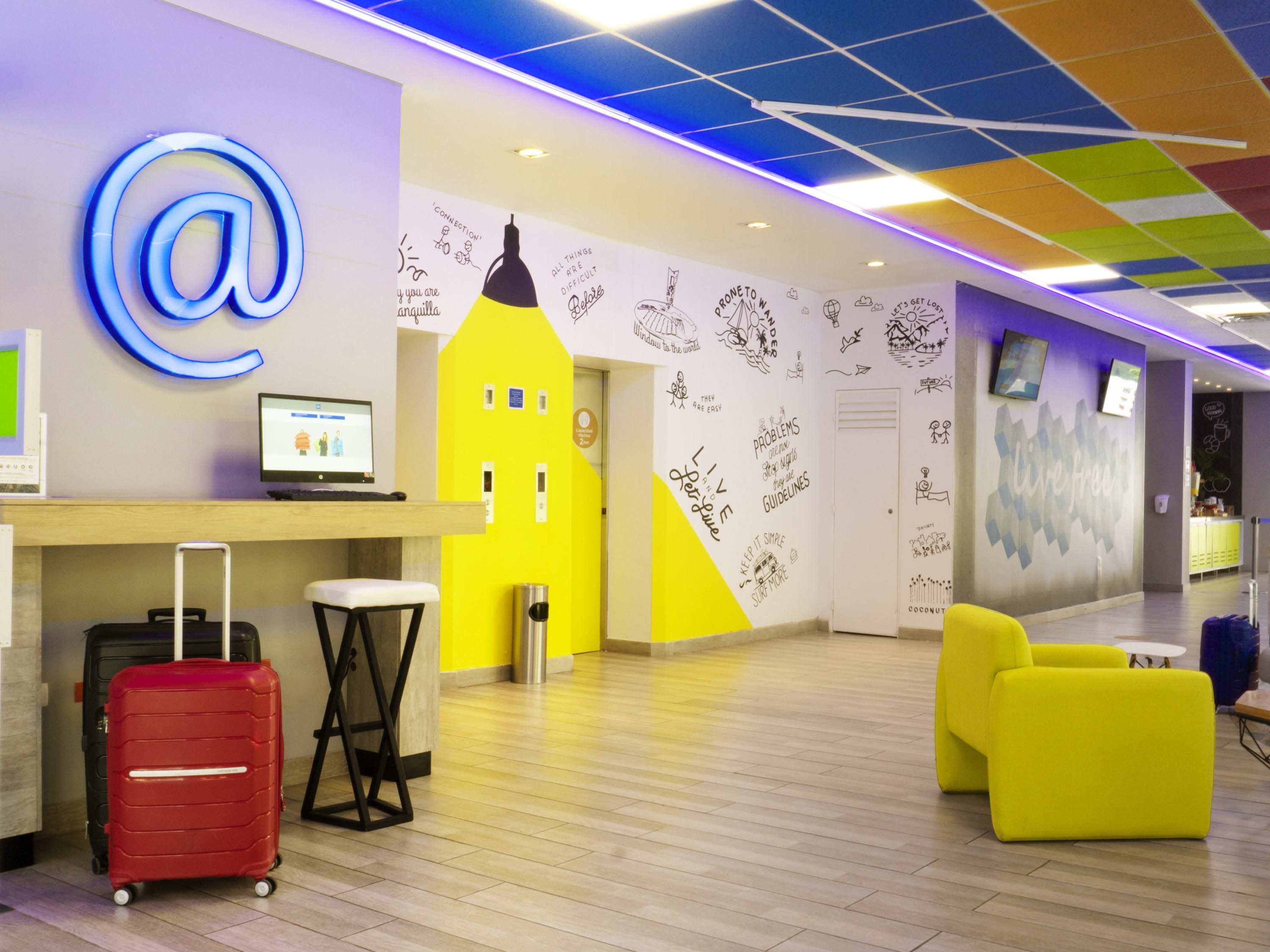 A modern hotel lobby with a large '@' sign, a computer, a yellow wall, and a reception desk.