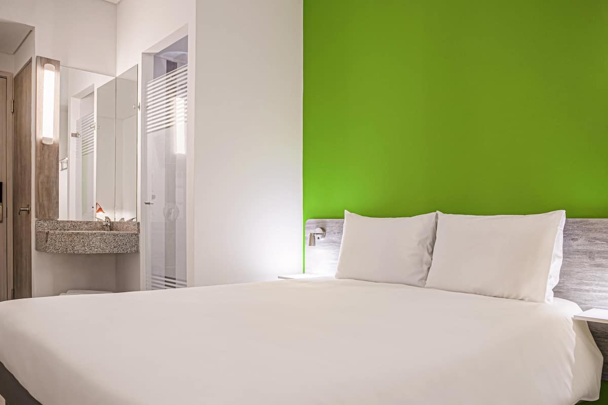 A modern hotel room with a large bed, a green accent wall, and a bathroom with a glass shower enclosure.