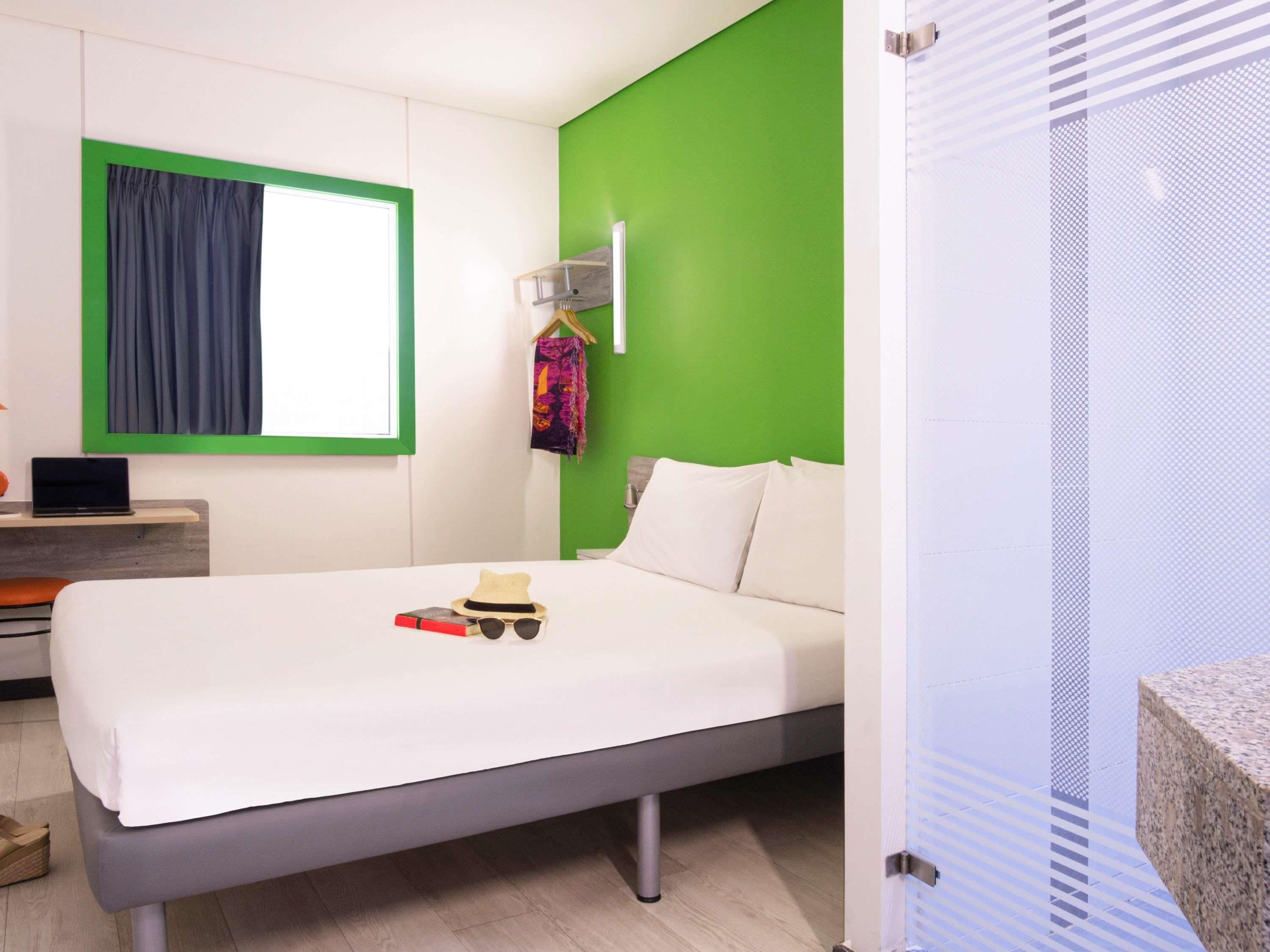 A modern hotel room with a bed, a desk with a laptop, a green accent wall, and a window with curtains.