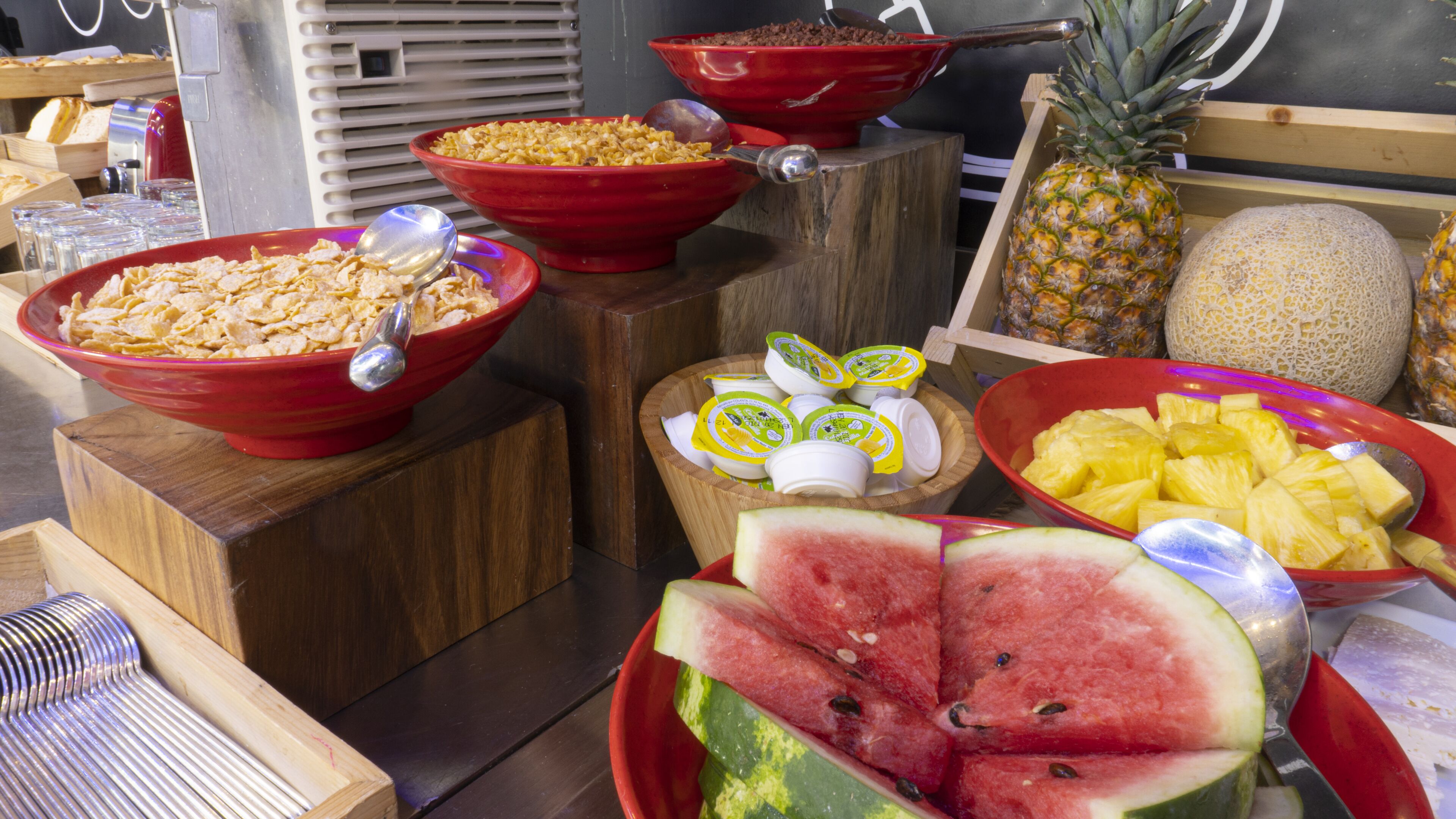 A breakfast buffet with various food items including cereal, yogurt, pineapple, watermelon, and melon.