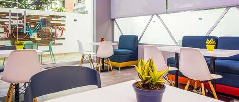 Un espacio interior moderno con mesas y sillas, una bicicleta decorativa, y un amplio ventanal con vistas a edificios y árboles.