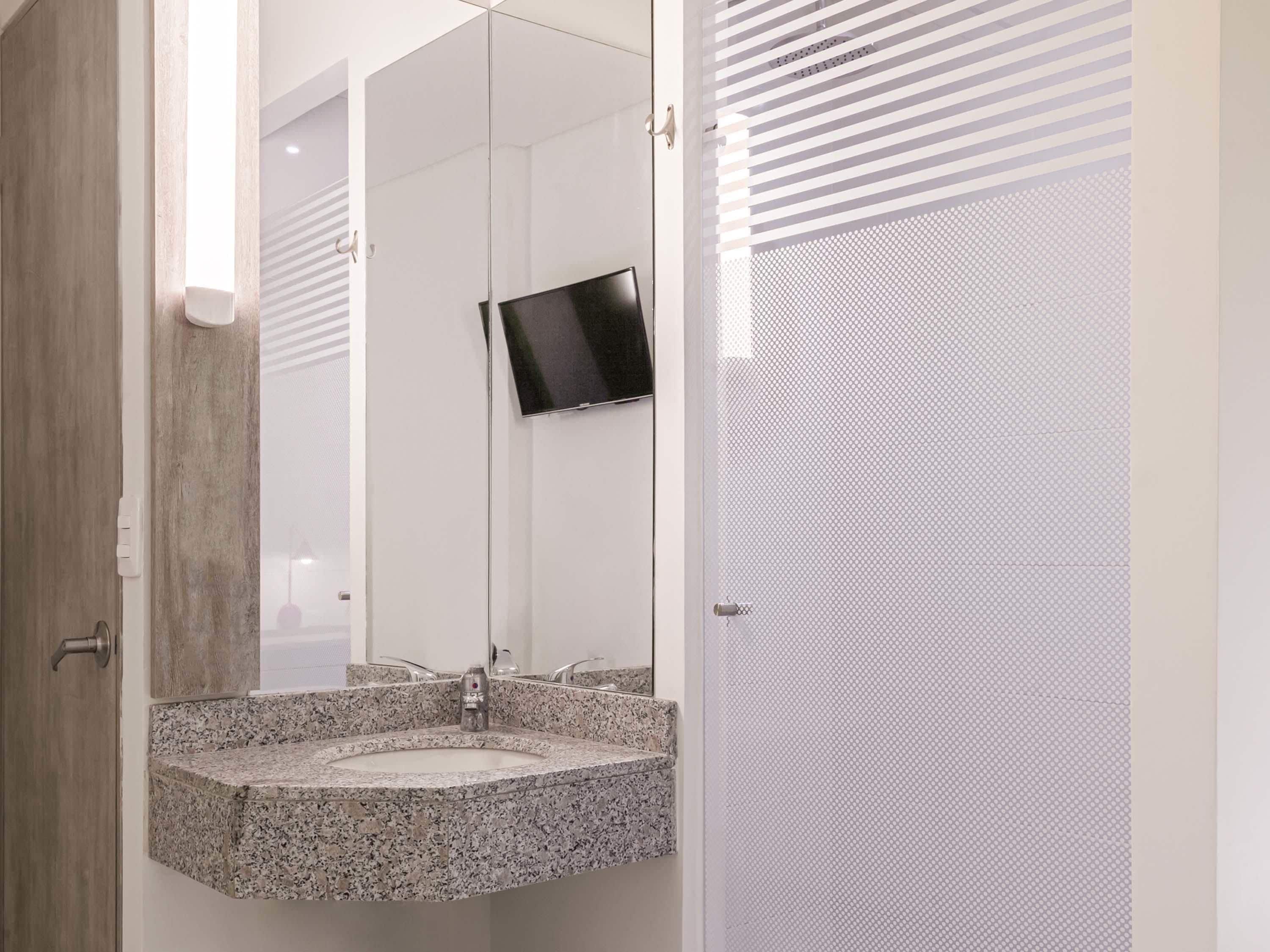 A bathroom with a granite countertop sink, a mirror, and a flat-screen TV mounted on the wall.