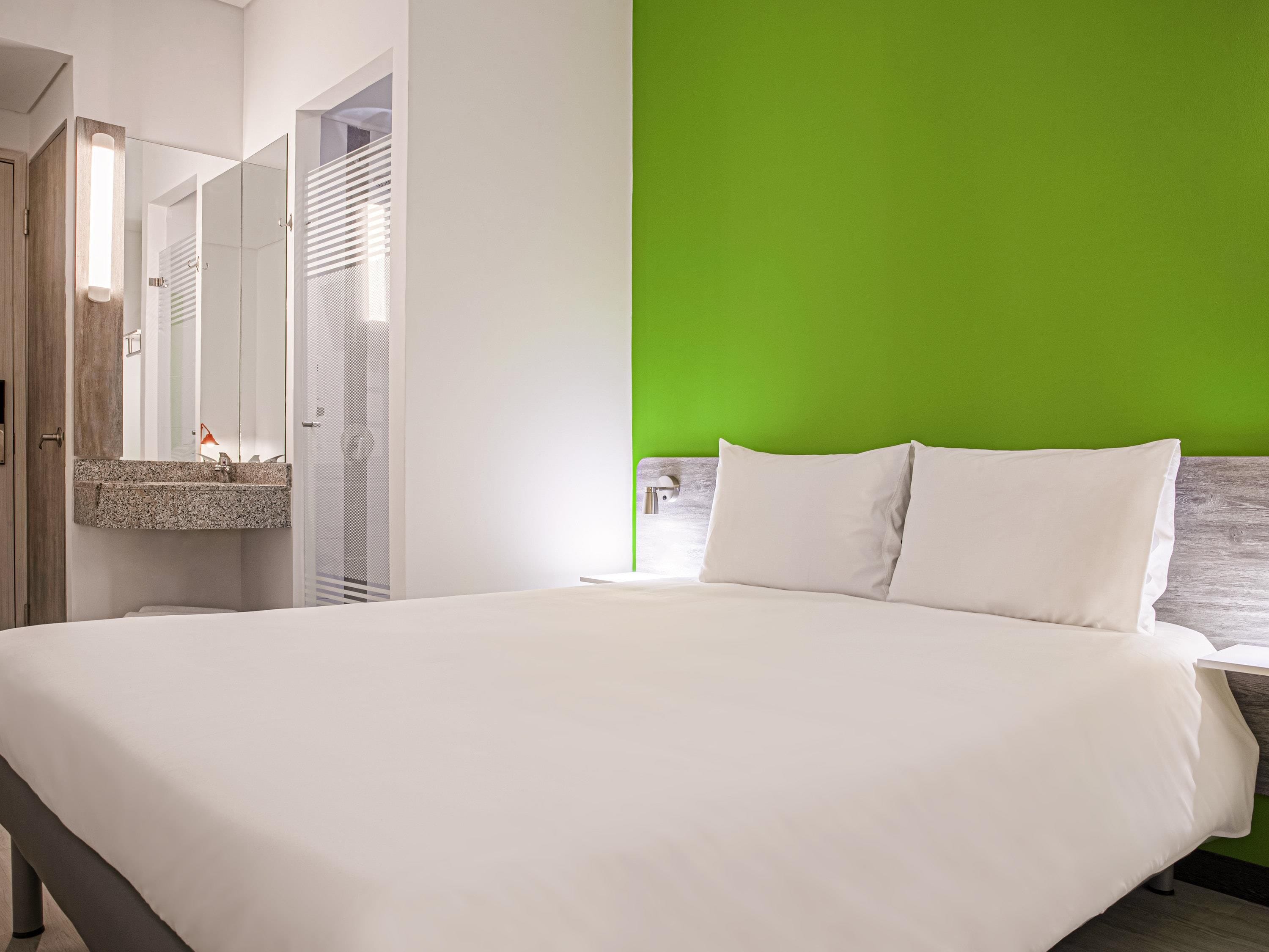 A modern hotel room with a large bed, a green accent wall, and a bathroom with a glass shower enclosure.